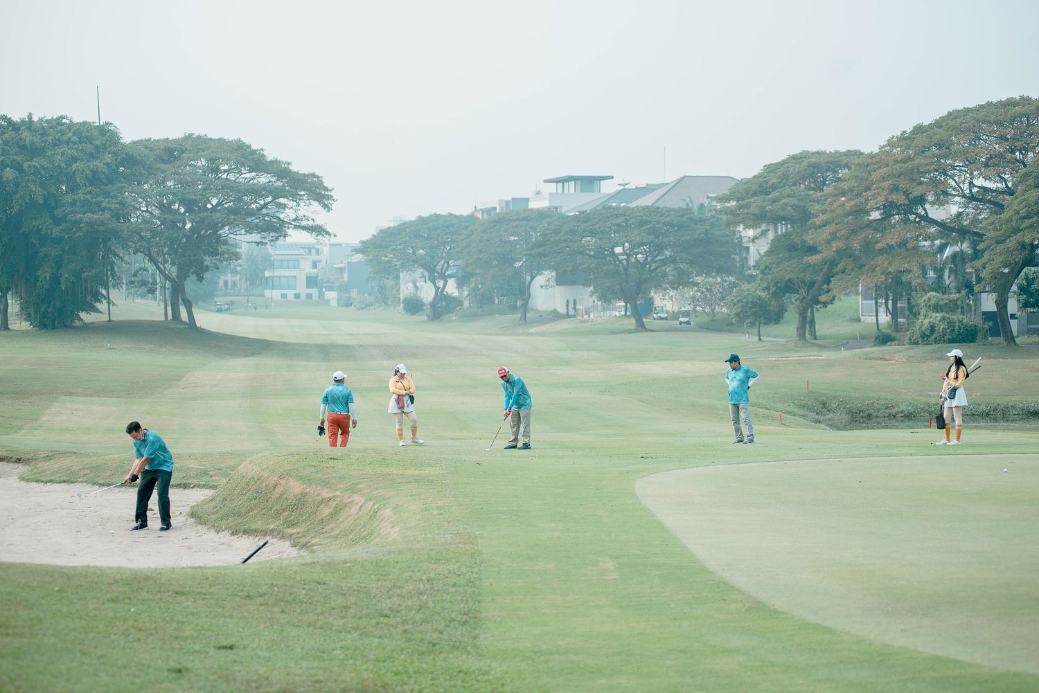 Golfers playing on a misty course surrounded by large tropical trees.