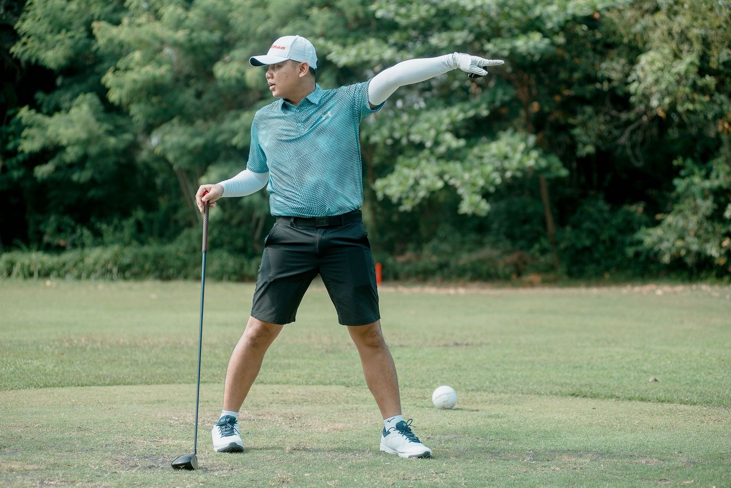 Golfer in light blue polo and dark shorts demonstrates stretching technique on golf course fairway.