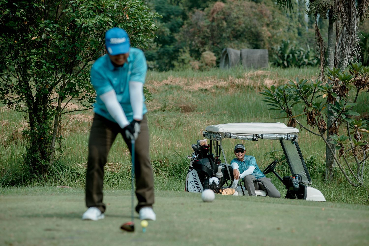Golfer in blue attire prepares to putt while golf cart with players waits in background.