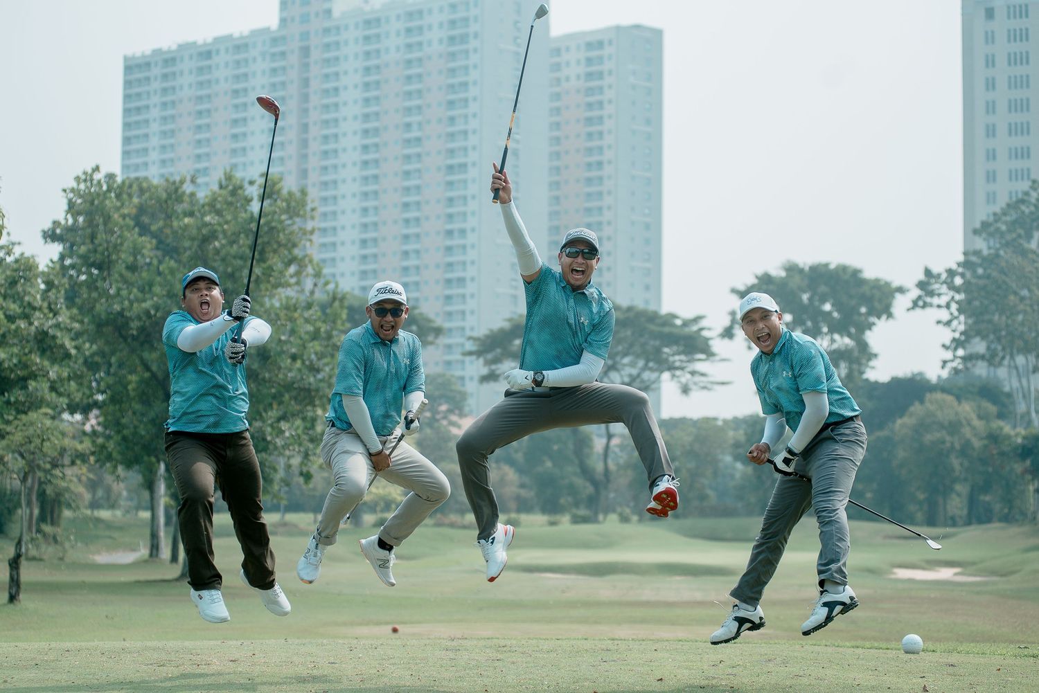 Four golfers in matching turquoise shirts celebrate with jumps and poses on a golf course with high-rise buildings in background.
