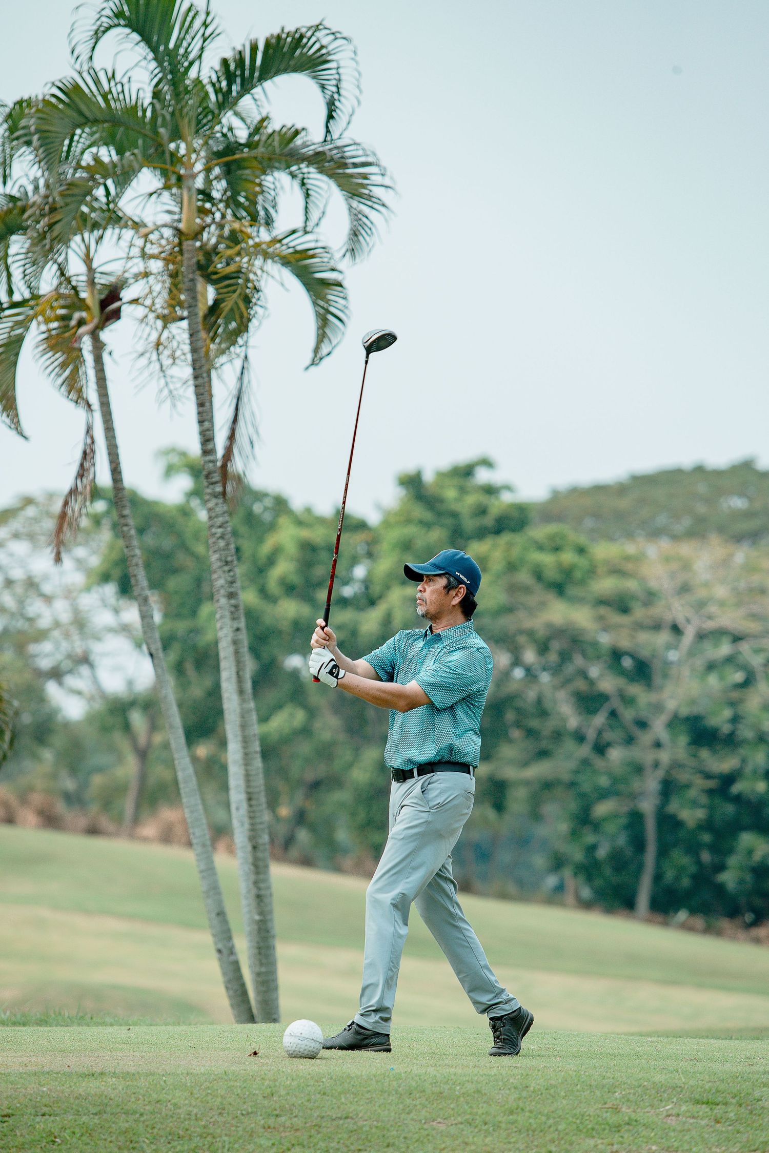 Golfer in teal shirt and khaki pants takes a swing on tropical course with palm trees in background.