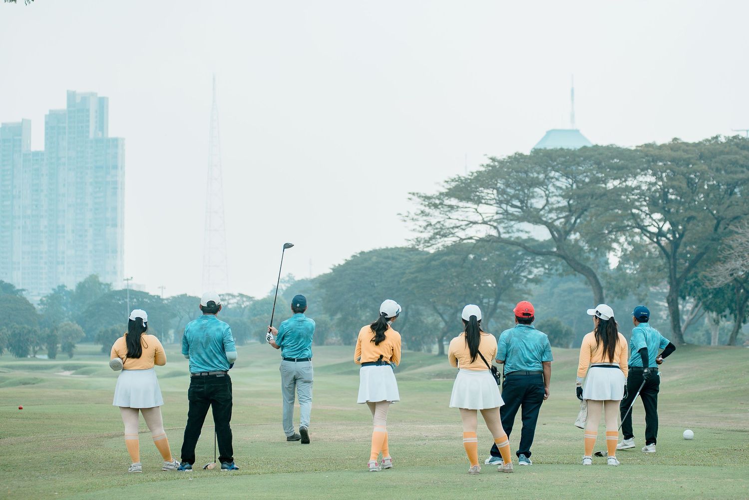 Group of people practicing golf swings on a grassy field with tall trees and urban buildings in the background.
