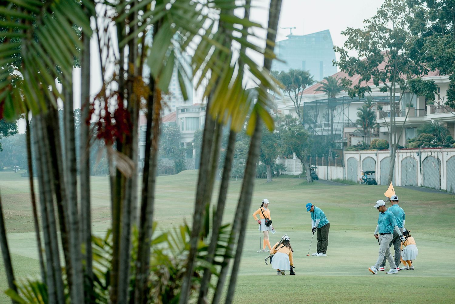 People playing golf on a lush green course with palm trees and buildings in the background on a cloudy day.
