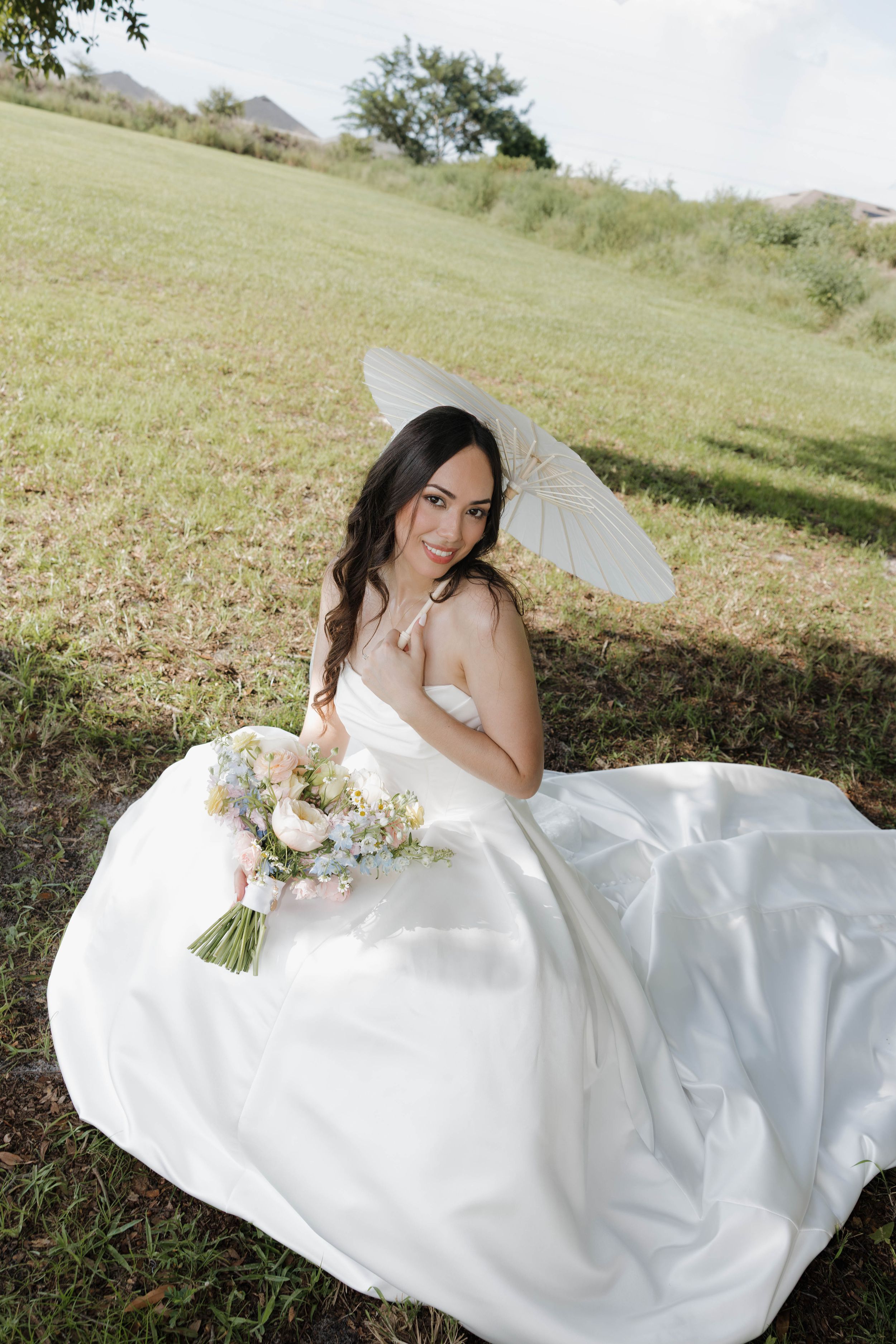 Bride in white gown sits on grass holding floral bouquet and white parasol umbrella, smiling outdoors.