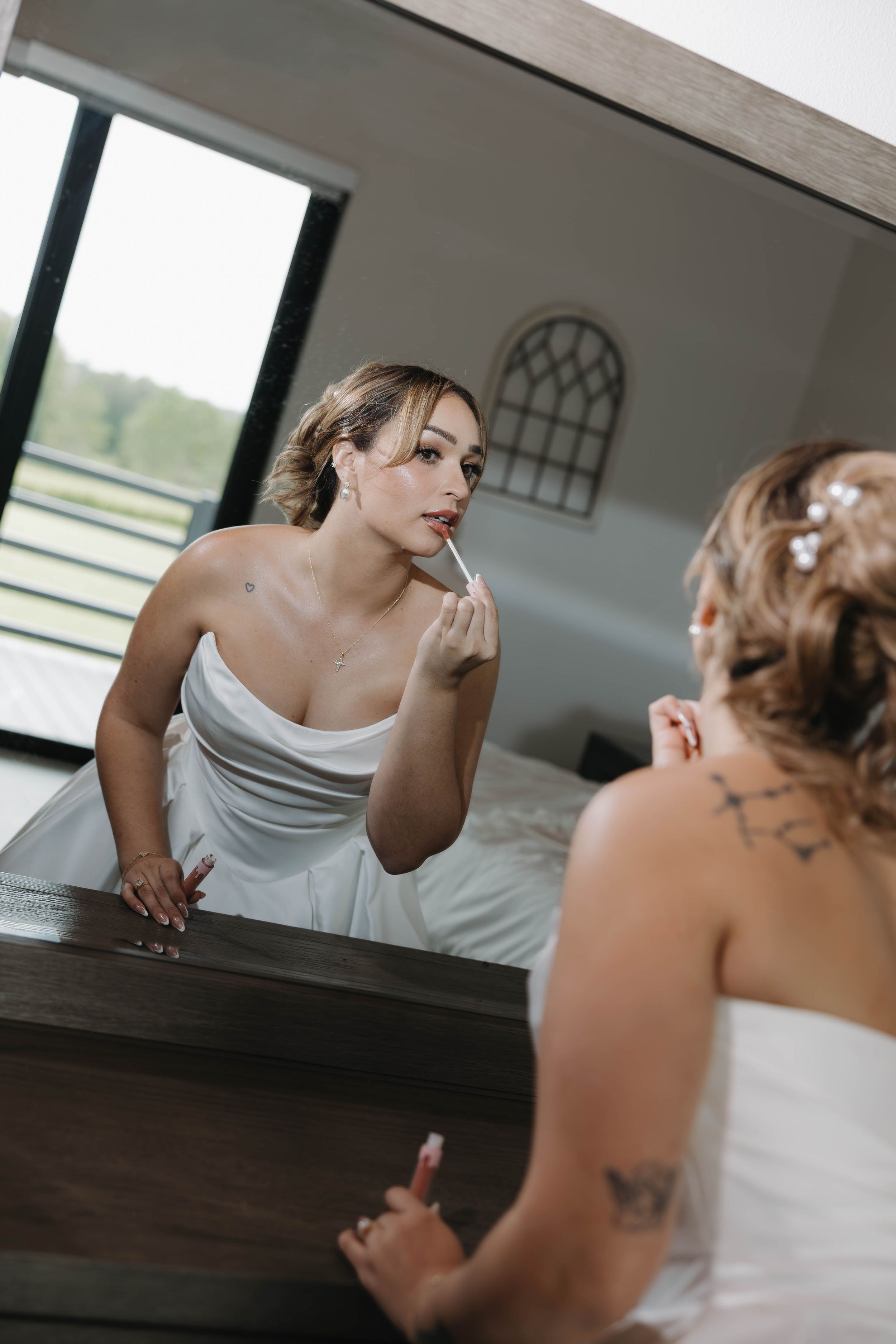 Bride in white strapless gown applying lipstick while looking in mirror, tattoo visible, hair accessories, wedding day preparation.