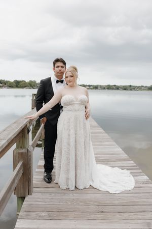 Bride and groom pose on a wooden dock by a lake, she in lace gown, he in black tuxedo.
