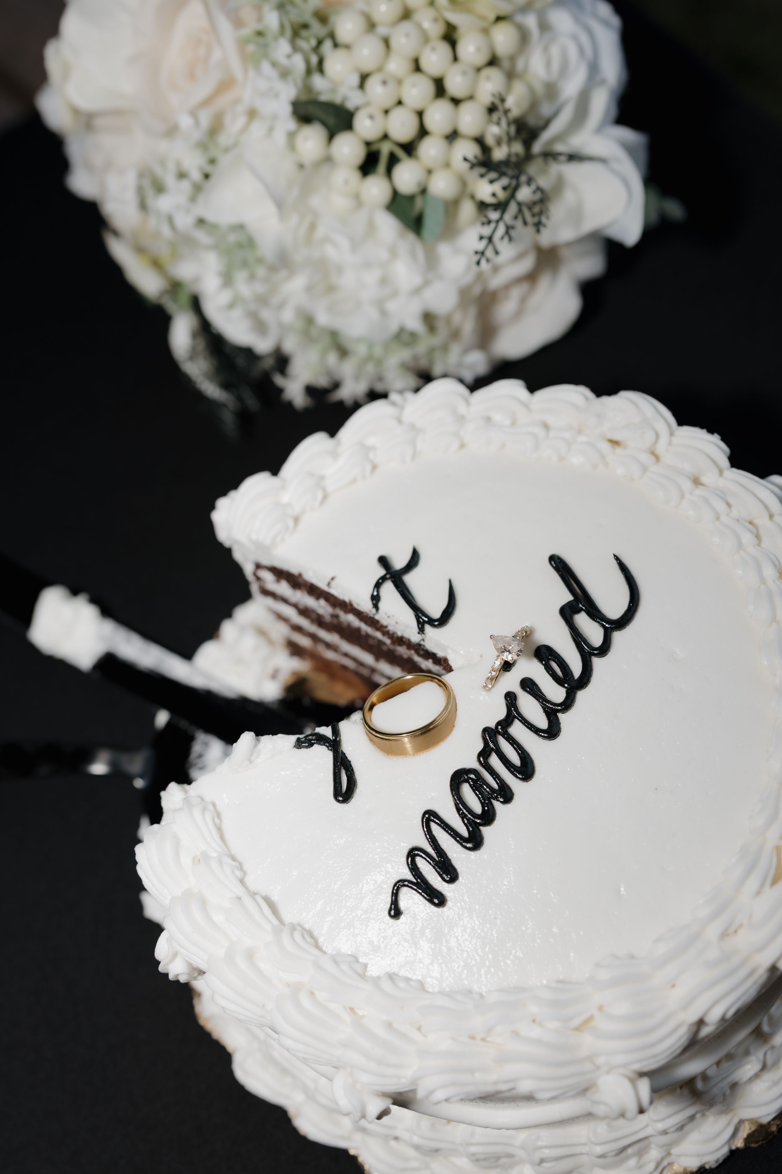 White wedding cake with 'just married' script, gold ring, and white floral bouquet on dark background.