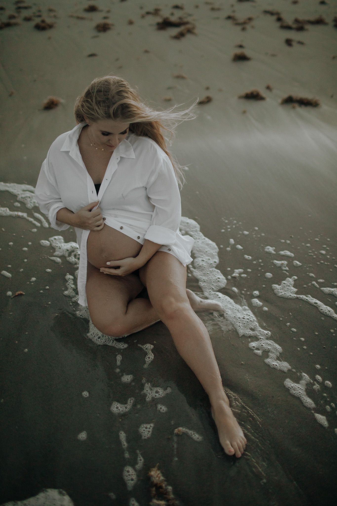 Beach maternity photos in a white shirt and black bikini sits in the warm ocean water on florida's treasure coast.