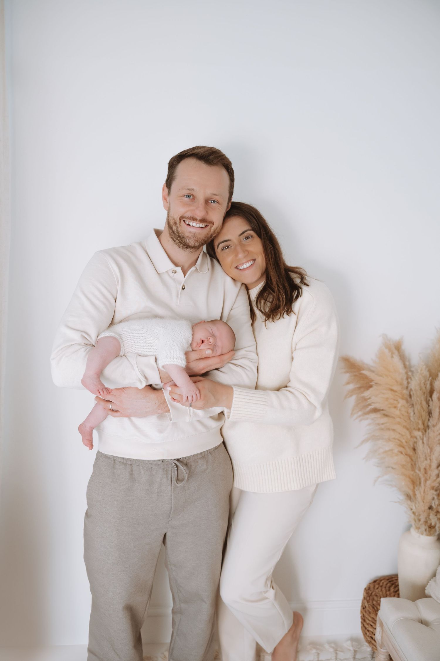 A family poses together in coordinated white and cream outfits against a neutral background with pampas grass decor.