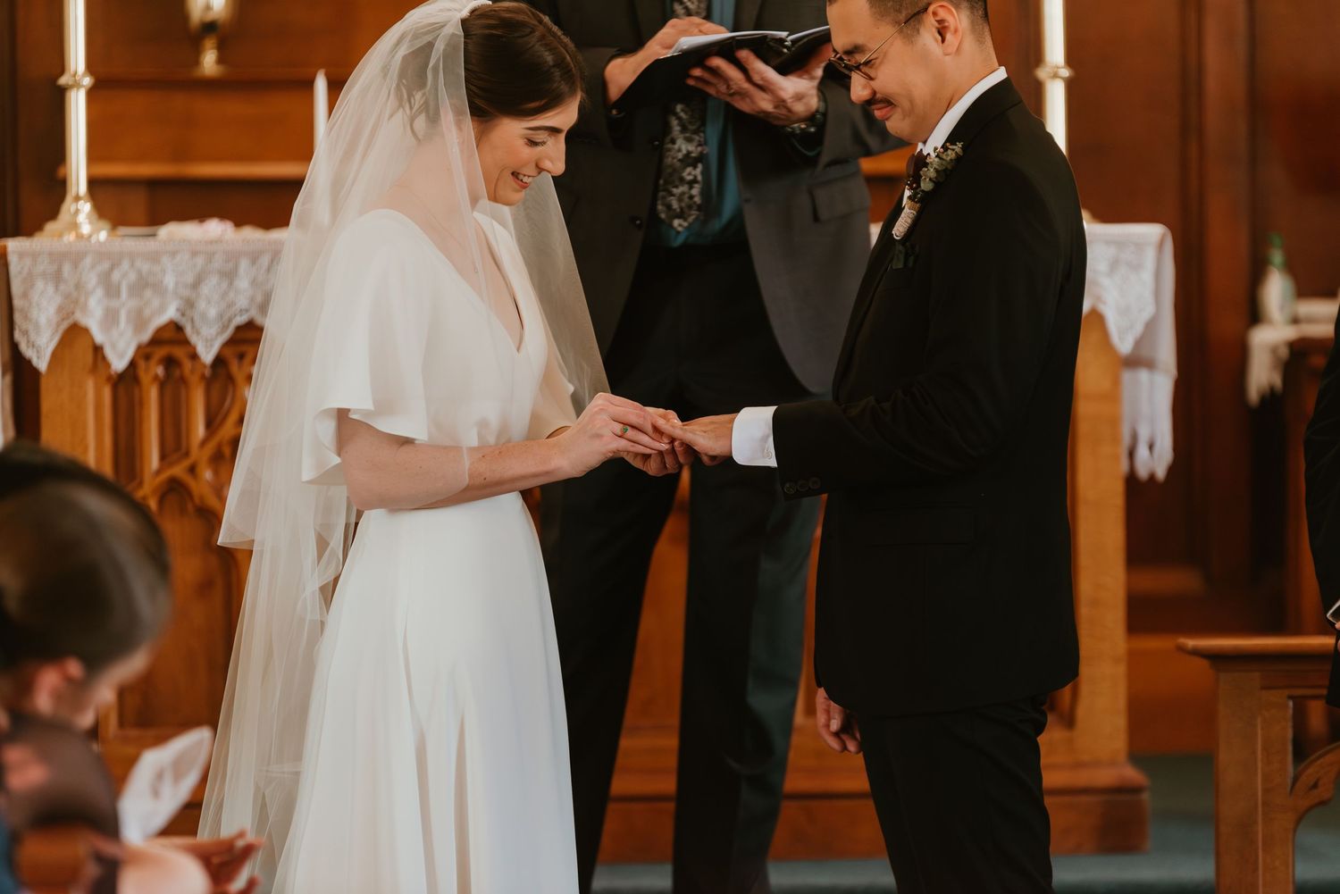 Bride and groom exchange rings during a church wedding ceremony, officiant reading from a book in the background.