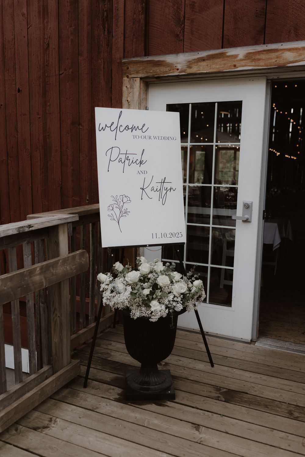 Wedding welcome sign and white floral arrangements displayed on rustic wooden deck against barn wood siding.