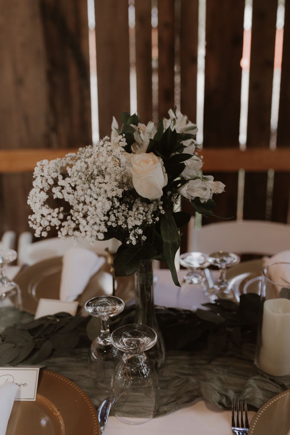 Elegant wedding reception table setting with white floral centerpieces and glassware in a rustic wooden venue.