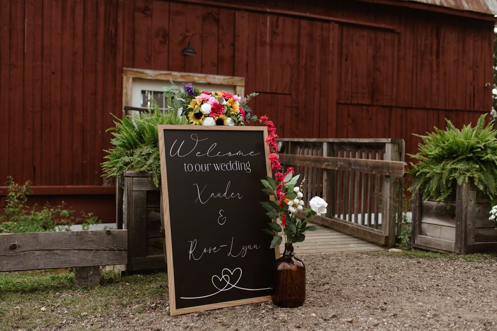A rustic red barn with a decorative chalkboard sign and floral arrangements creates a charming wedding venue entrance.