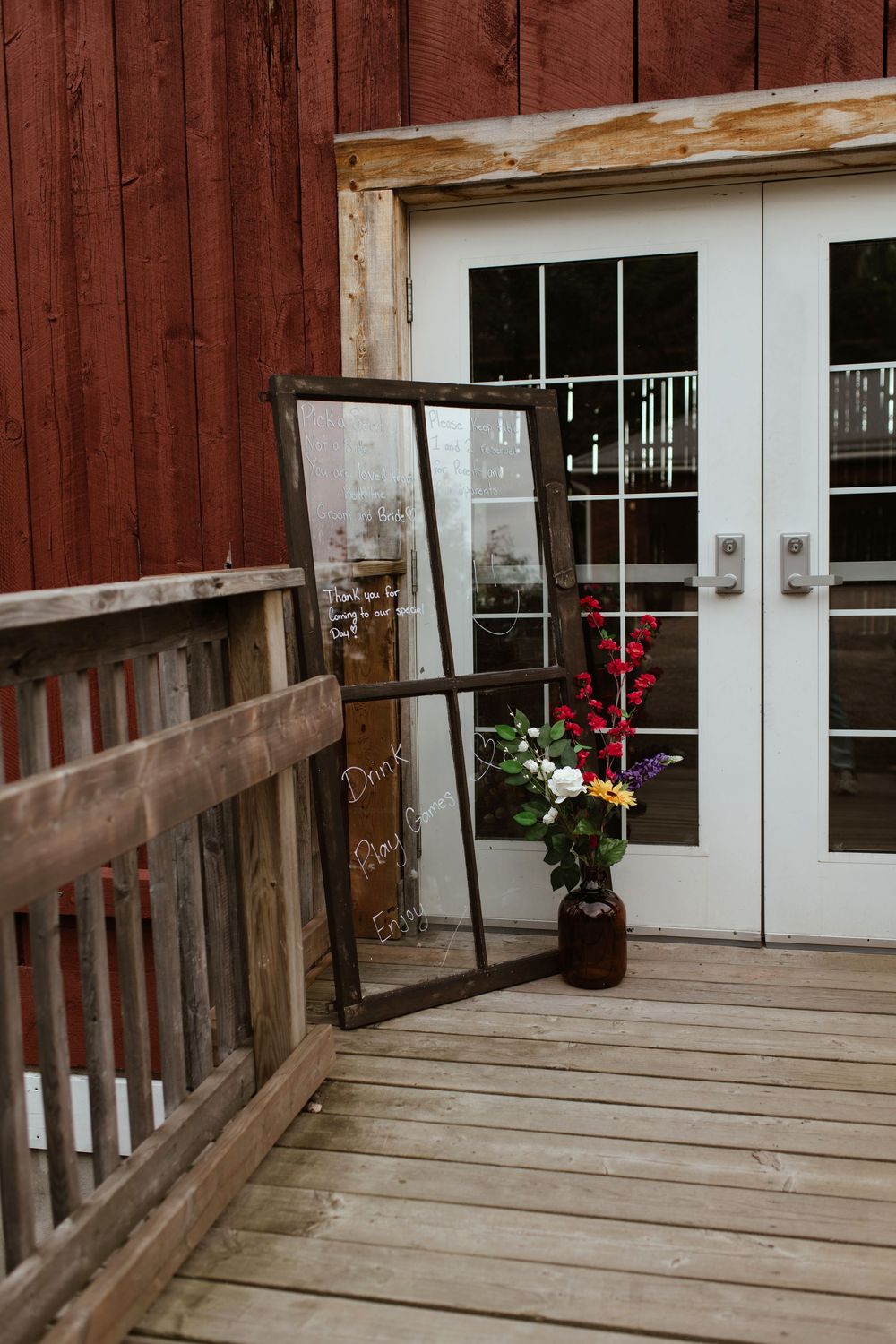 Rustic wooden deck entrance with red barn siding and white doors features decorative red flowers near the doorway.