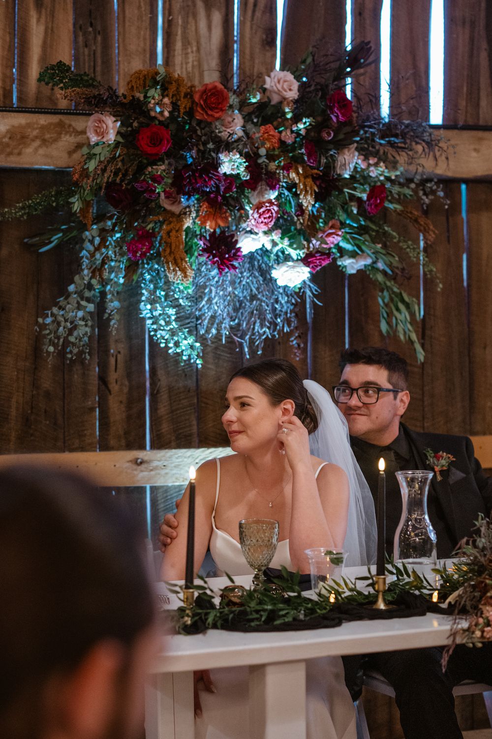Wedding couple seated at decorated head table with floral arrangements and hanging decor in rustic barn venue.