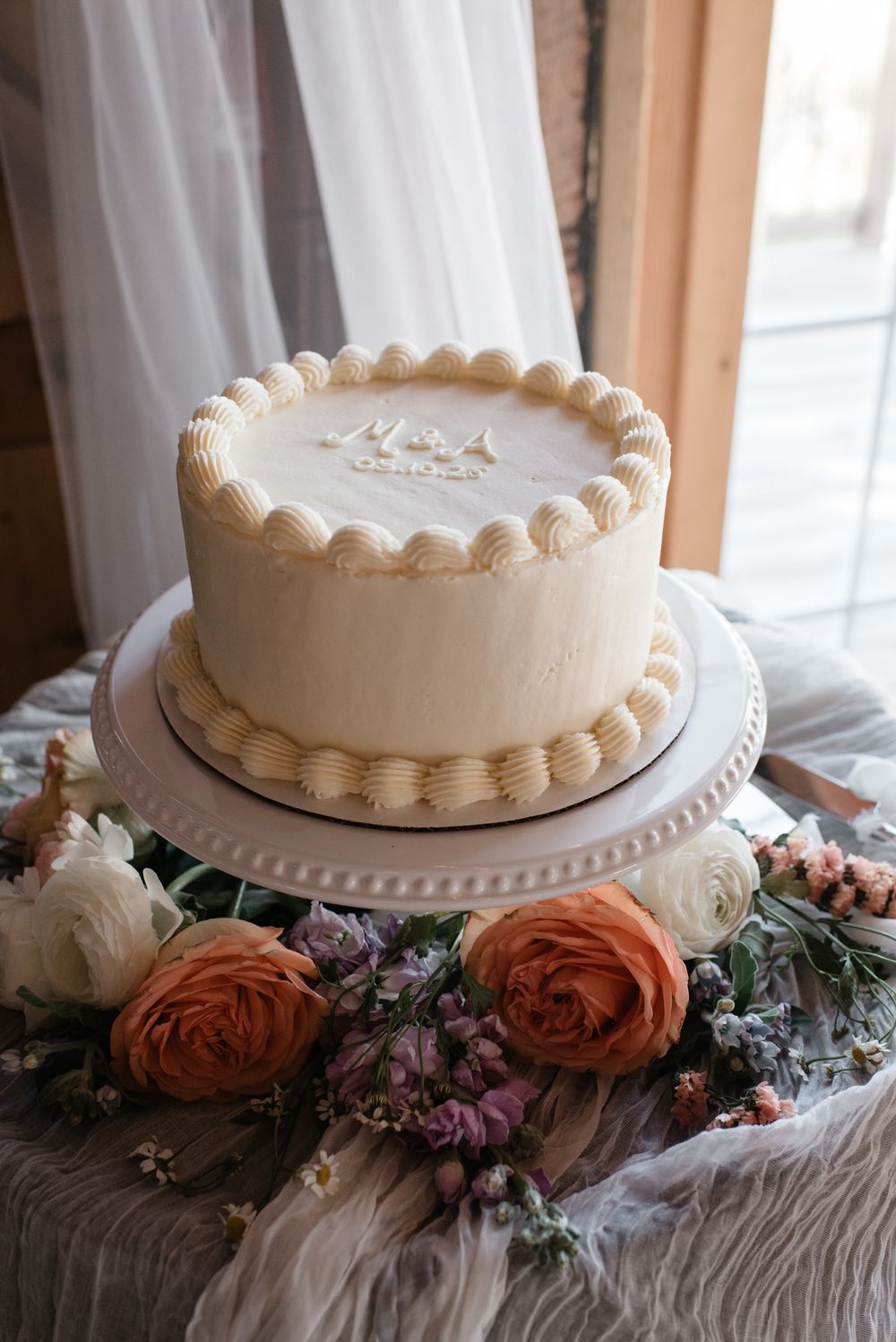 White wedding cake decorated with delicate piping sits atop a table adorned with coral roses and purple florals.