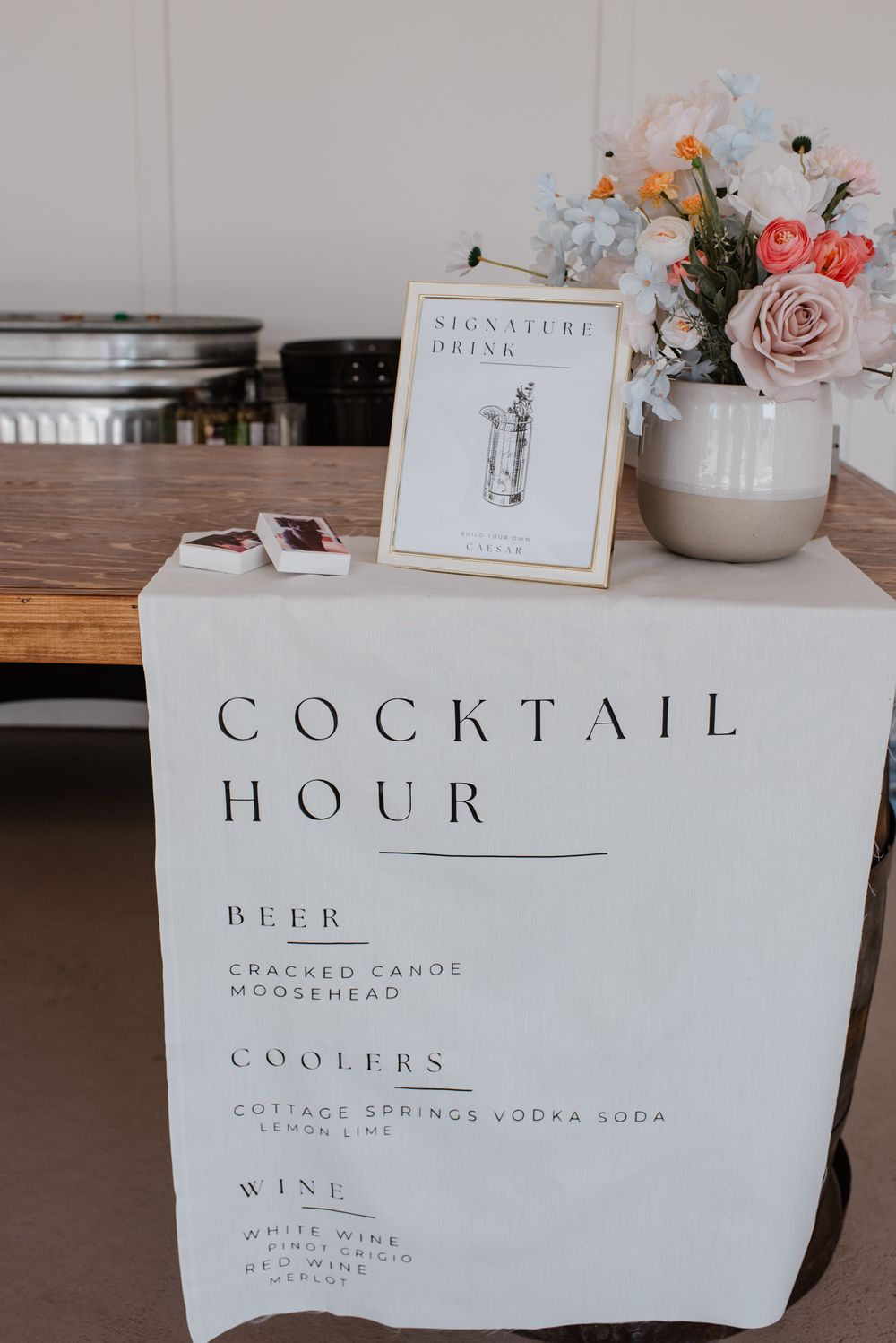 Minimalist white wedding bar menu signs displaying cocktail hour and drink options on wooden tables.