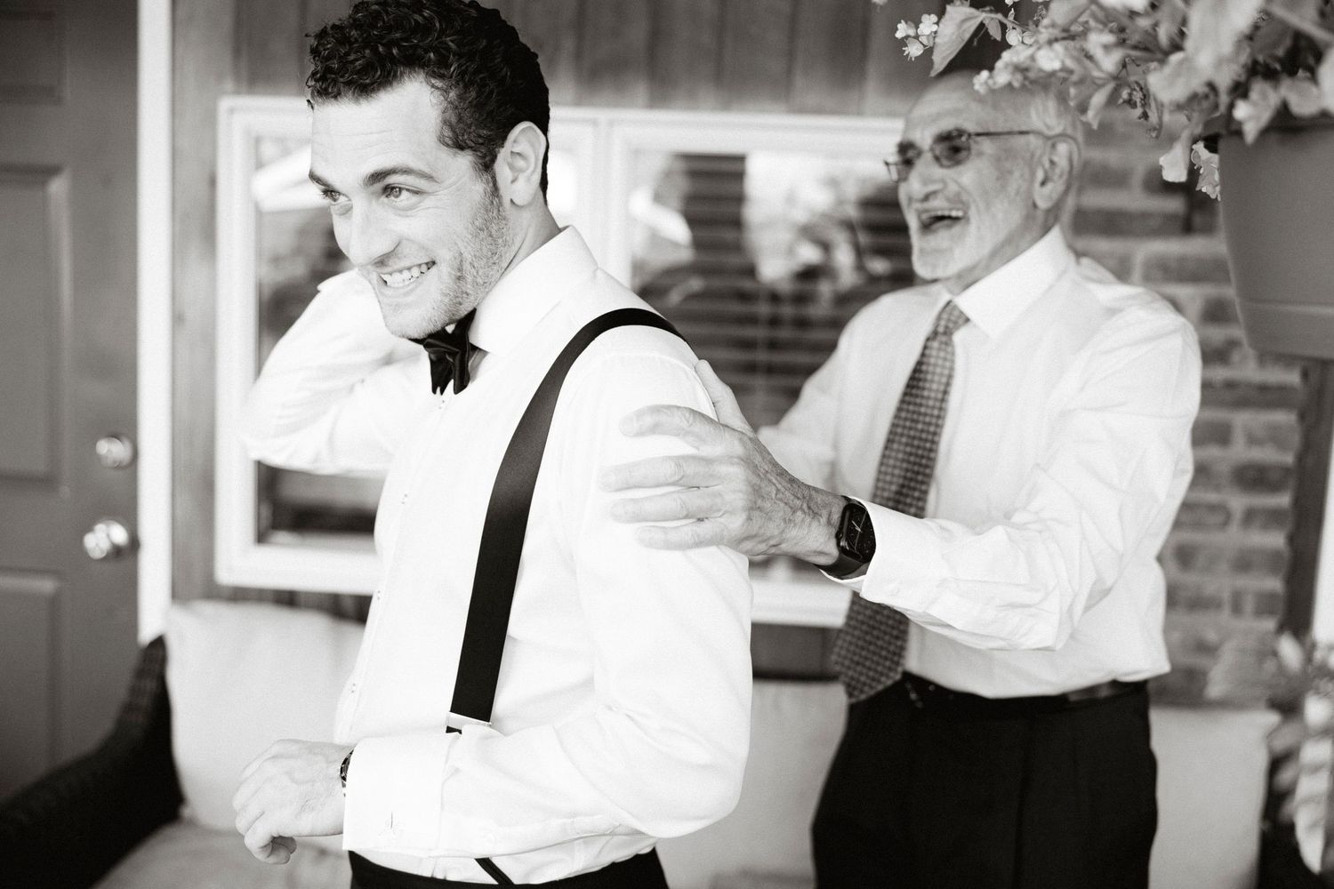 Black and white photo of two people getting ready for a formal event, with one adjusting the others suspenders.