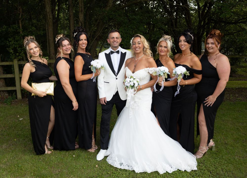 Wedding party poses outdoors in formal black and white attire for group photos in garden setting.
