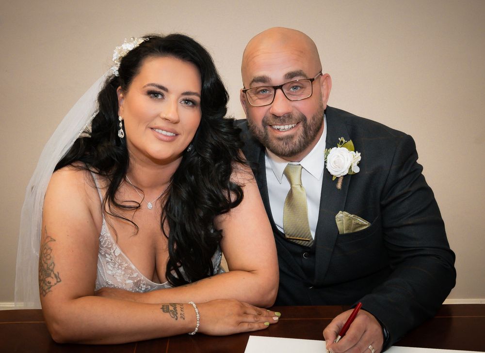 A couple poses together in formal wedding attire with a white boutonniere and yellow tie.