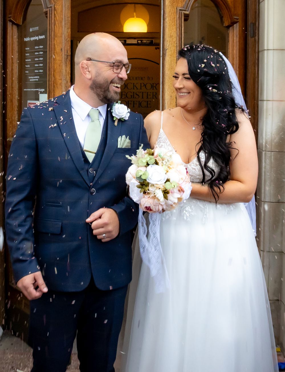 Happy newlyweds and wedding party celebrating with confetti outside a grand stone building entrance.
