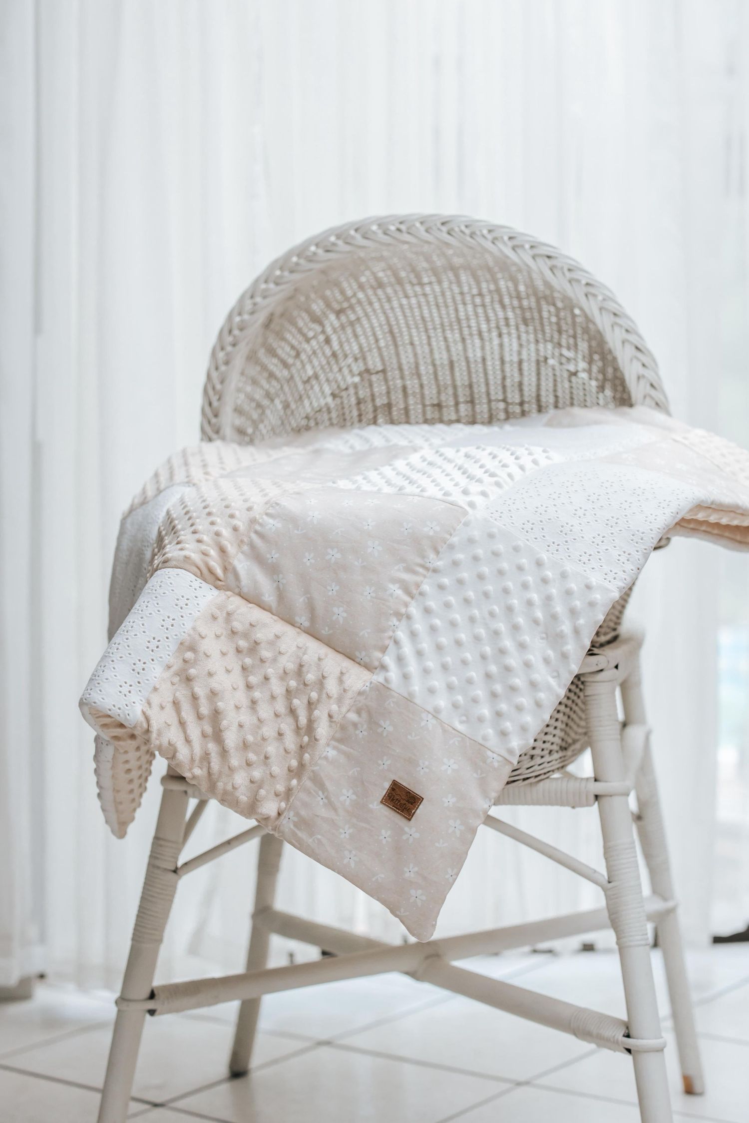 A white wicker chair with a cozy textured blanket draped over it sits against a light curtained background.
