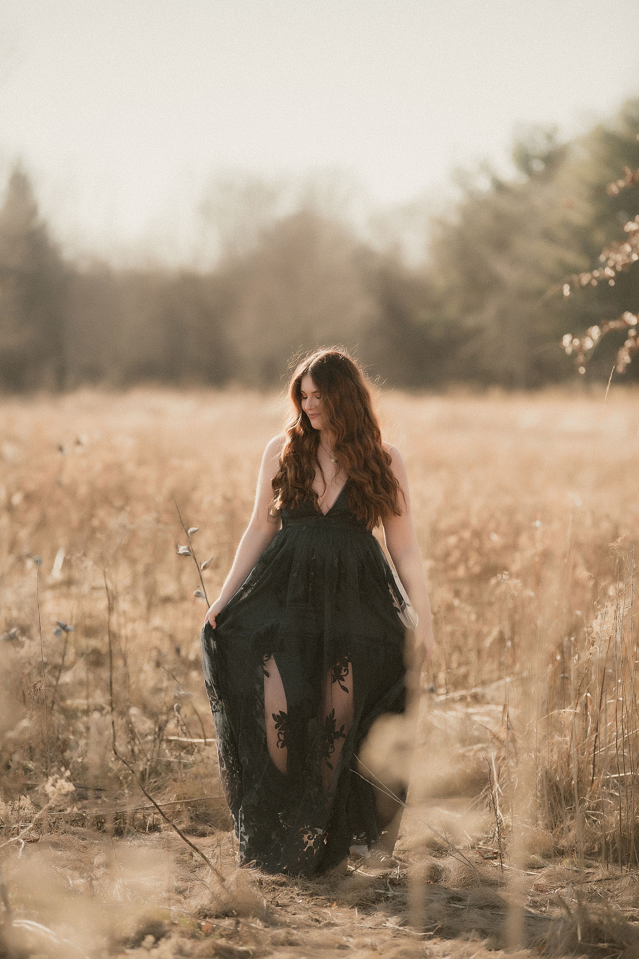 A person in a flowing black dress stands in a golden field at sunset with evergreen trees in the background.