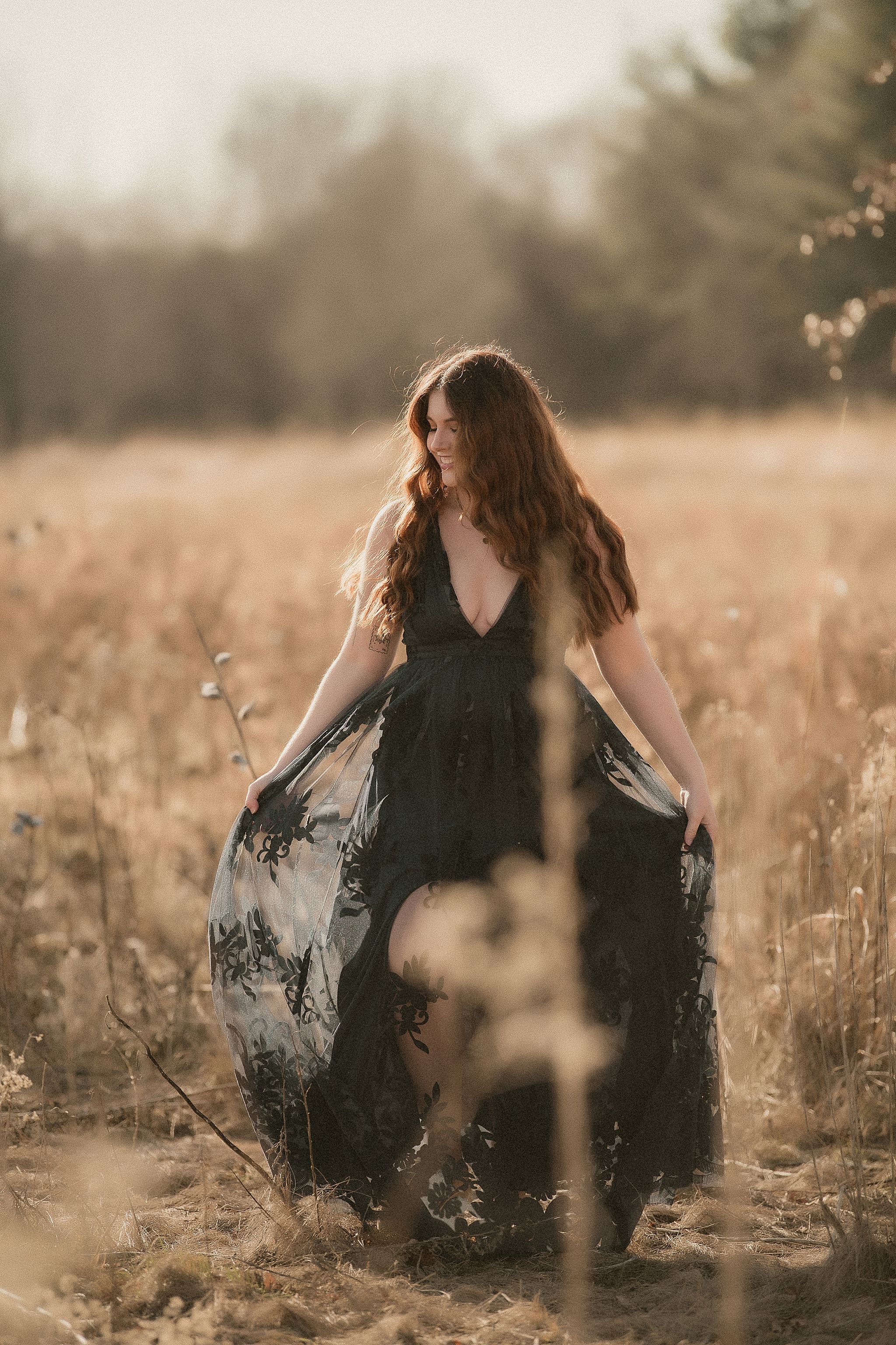 A person in a black dress sits gracefully in a sunlit meadow with tall grass and wildflowers at golden hour.