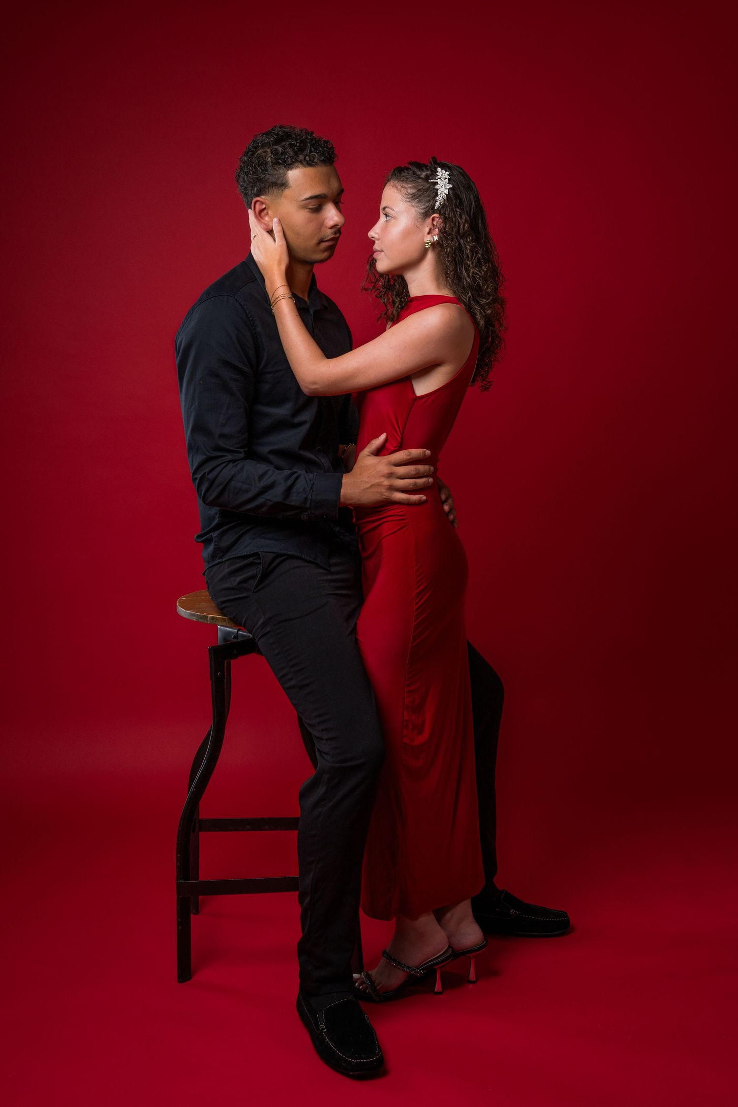 Romantic couple in black and red attire pose intimately against dark red backdrop with dramatic studio lighting.