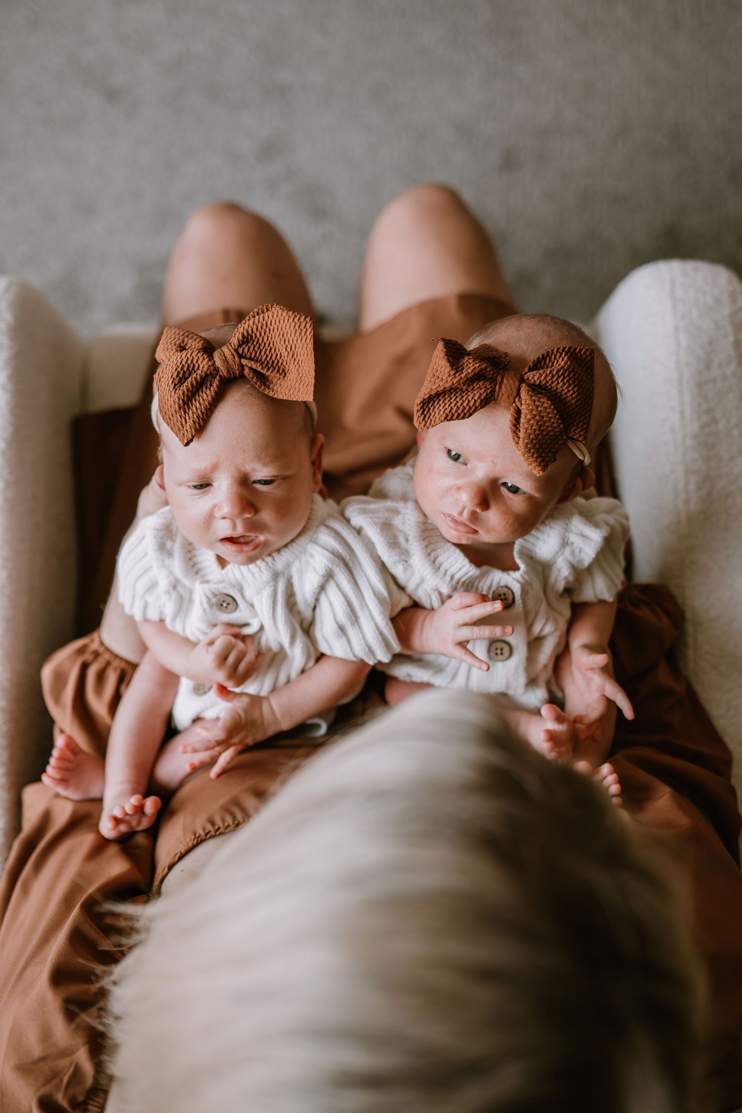Two sleeping infants in white outfits and brown bows lying together on a cozy beige blanket.