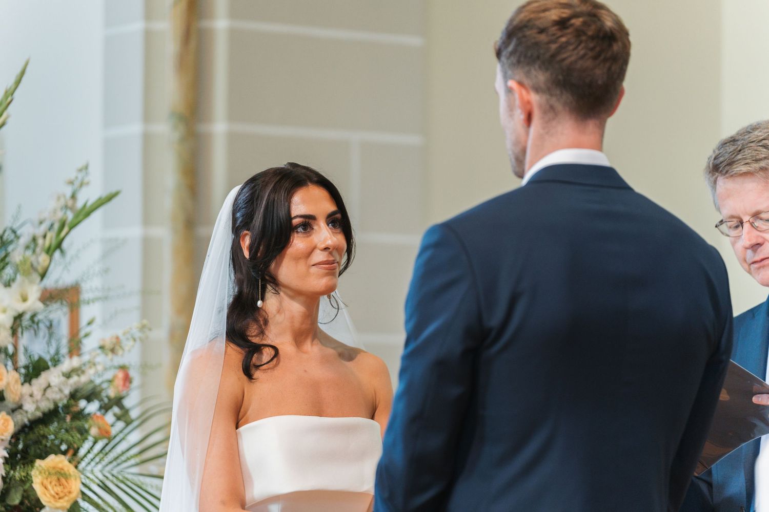 A couple exchanges vows during an elegant indoor wedding ceremony with greenery decorations and natural lighting.