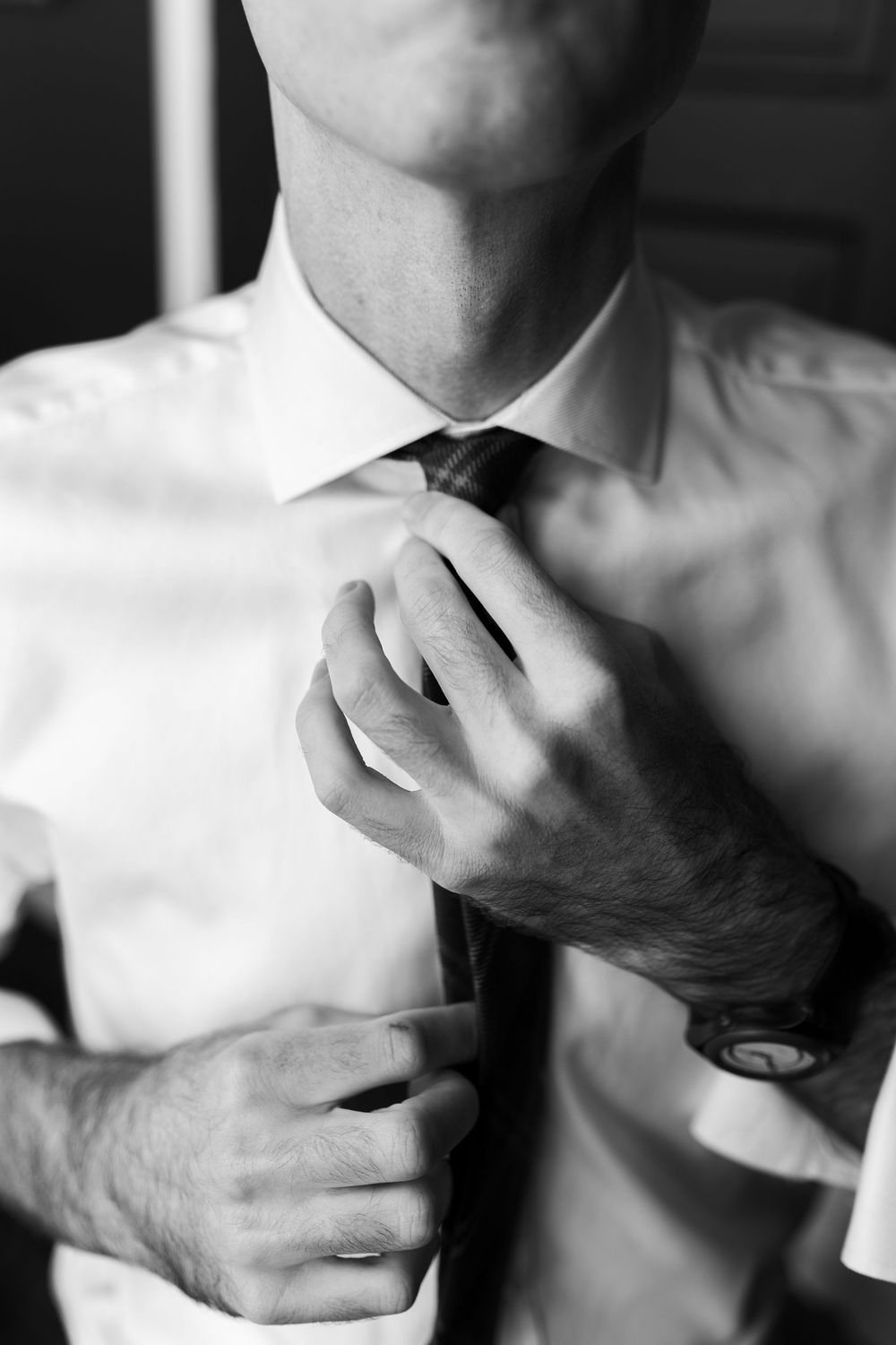 Black and white close-up of groom adjusting tie while getting ready for wedding.
