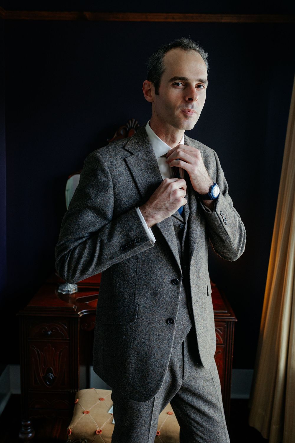 Dapper groom adjusting tie in grey tweed suit against a dark navy blue wall.