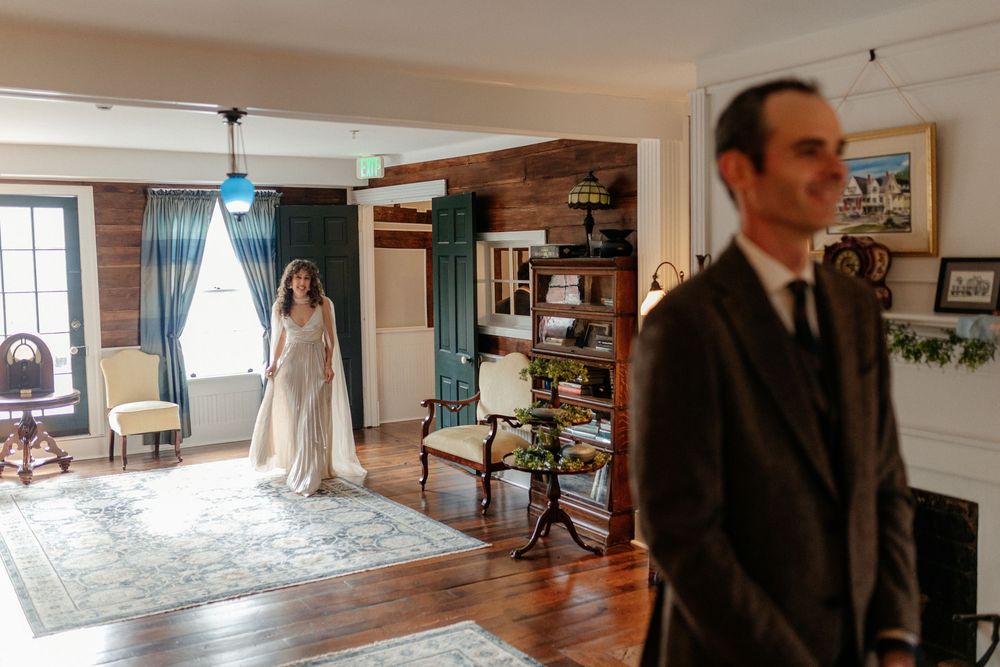Bride in flowing white gown approaches groom for first look in rustic wood-paneled room with vintage furnishings.