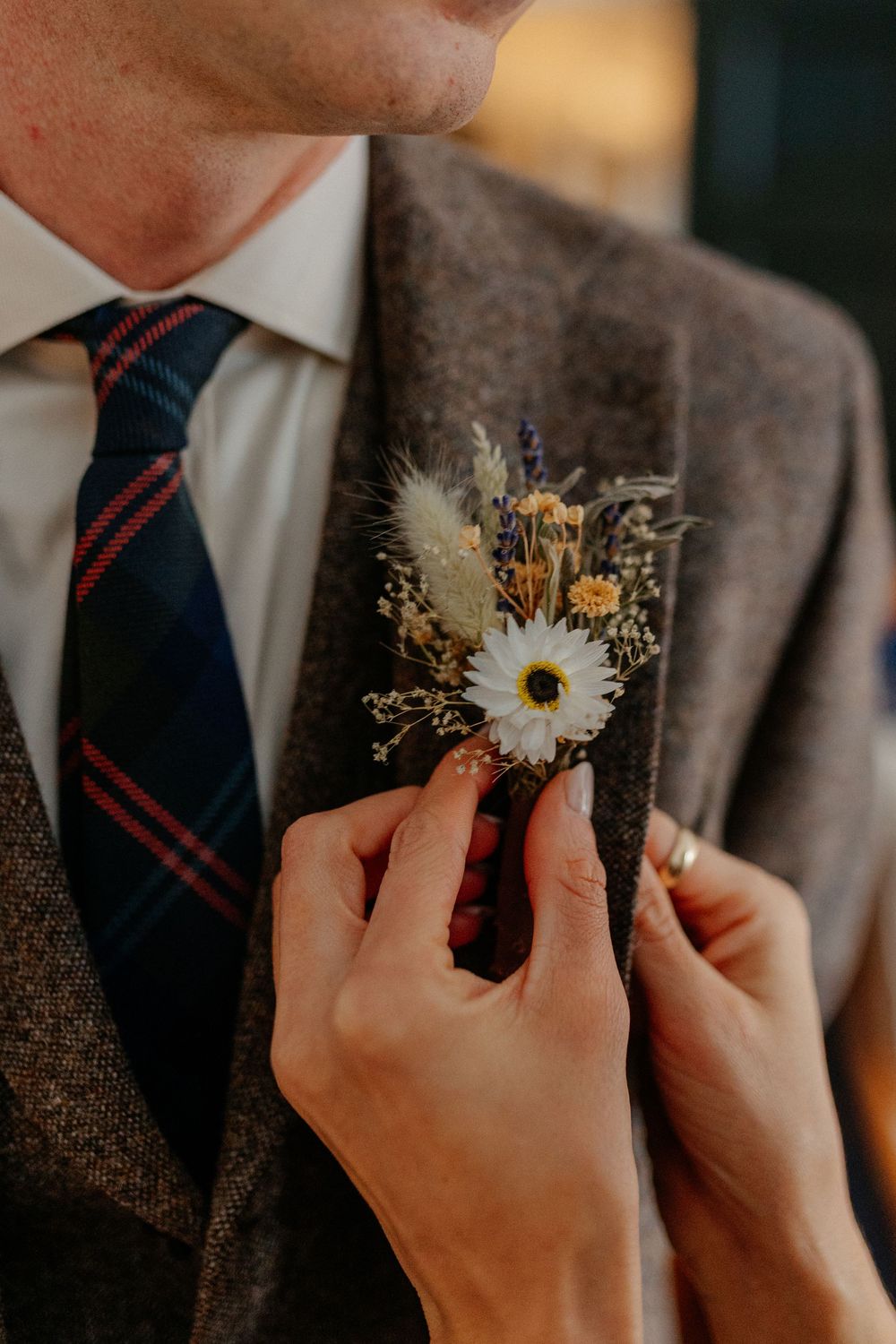 Hands pin a wildflower boutonniere with daisy and dried grasses onto groom's brown tweed suit jacket.
