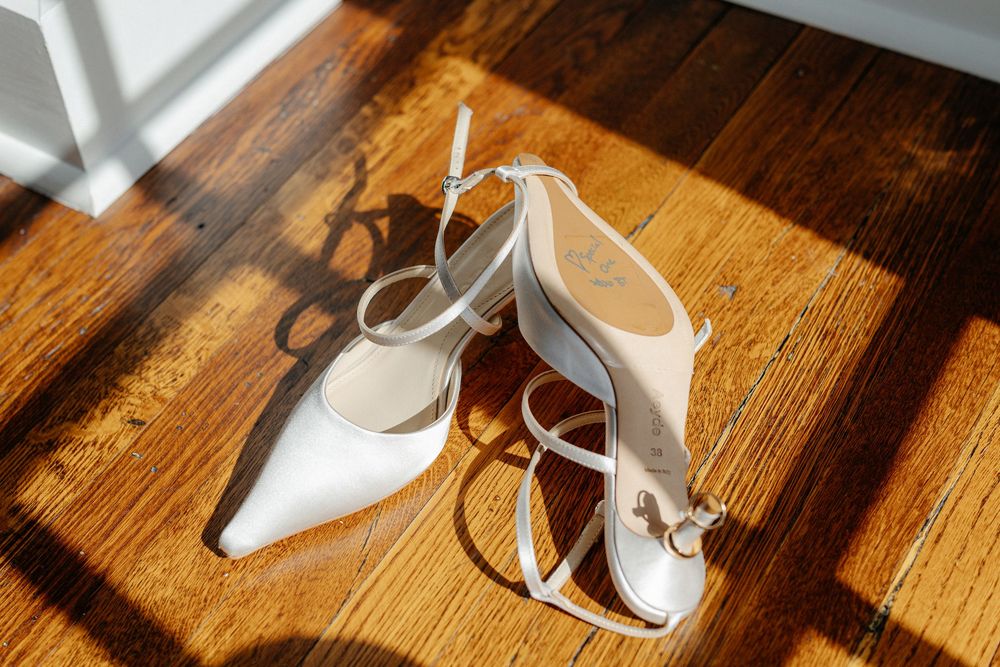 White bridal heels with ankle straps casting shadows on a sunlit hardwood floor.