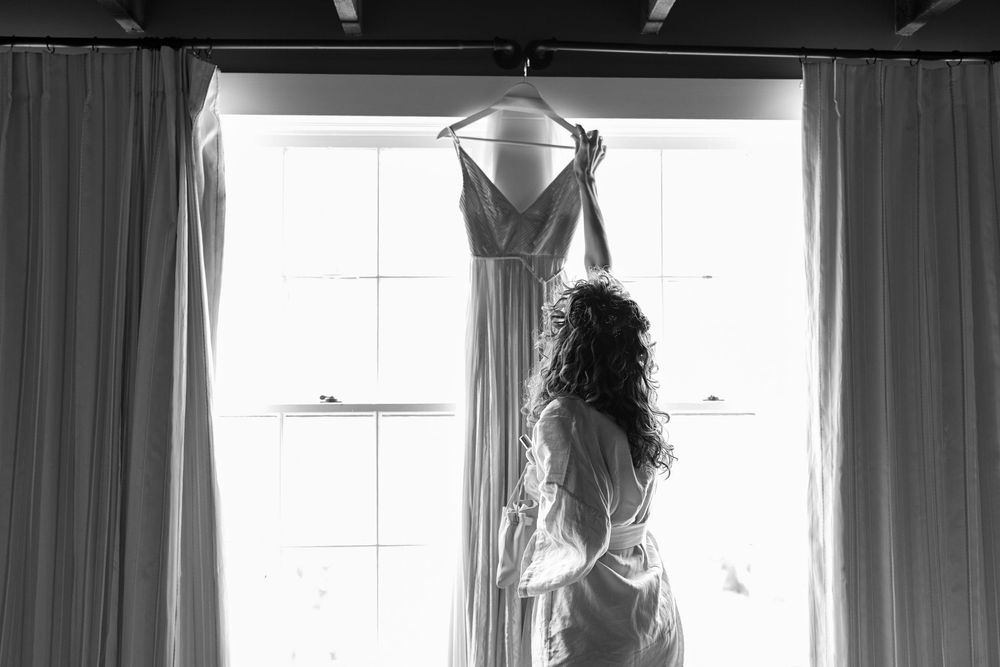 Black and white silhouette of bride holding wedding dress in front of bright window.