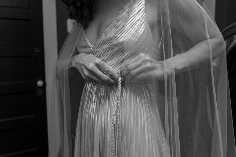 Bride adjusts braided belt on shimmering pleated wedding gown with sheer cape veil in black and white photo.