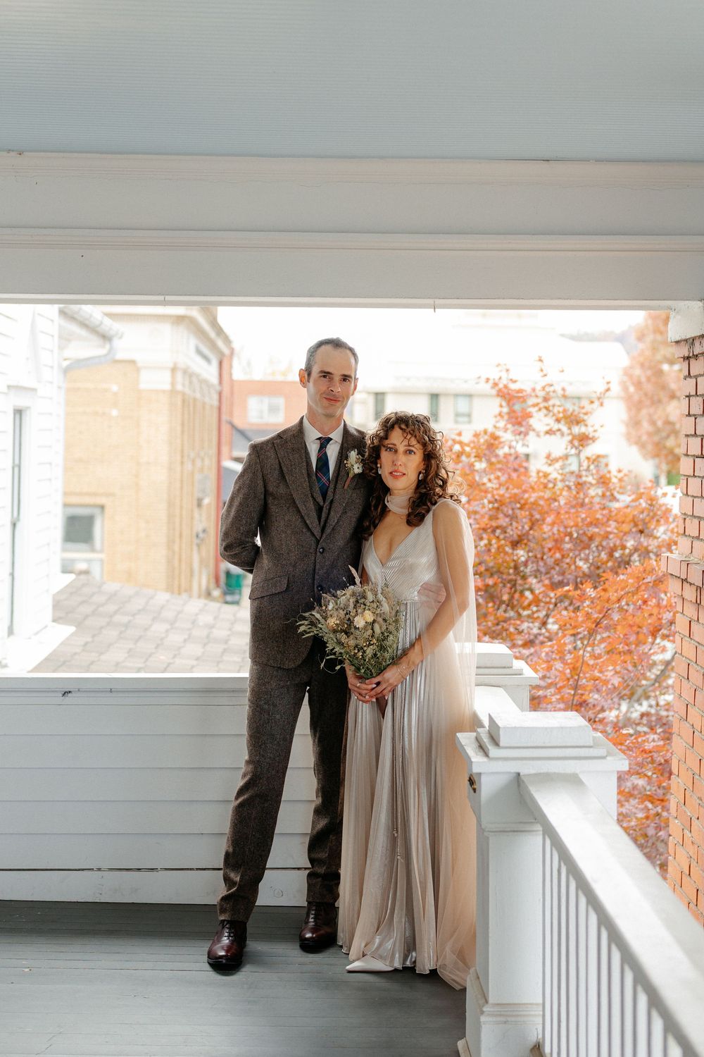 Bride and groom portrait on sunlit porch with autumn foliage, bride holding dried wildflower bouquet.