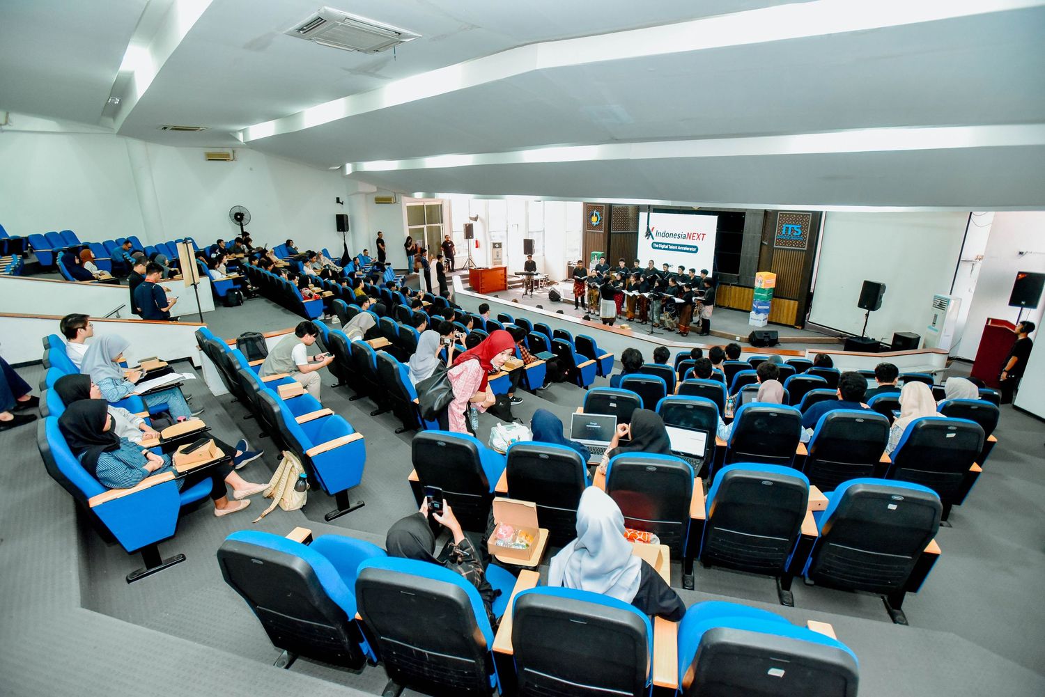 Students attend a lecture in a modern classroom with rows of blue chairs and computer workstations.