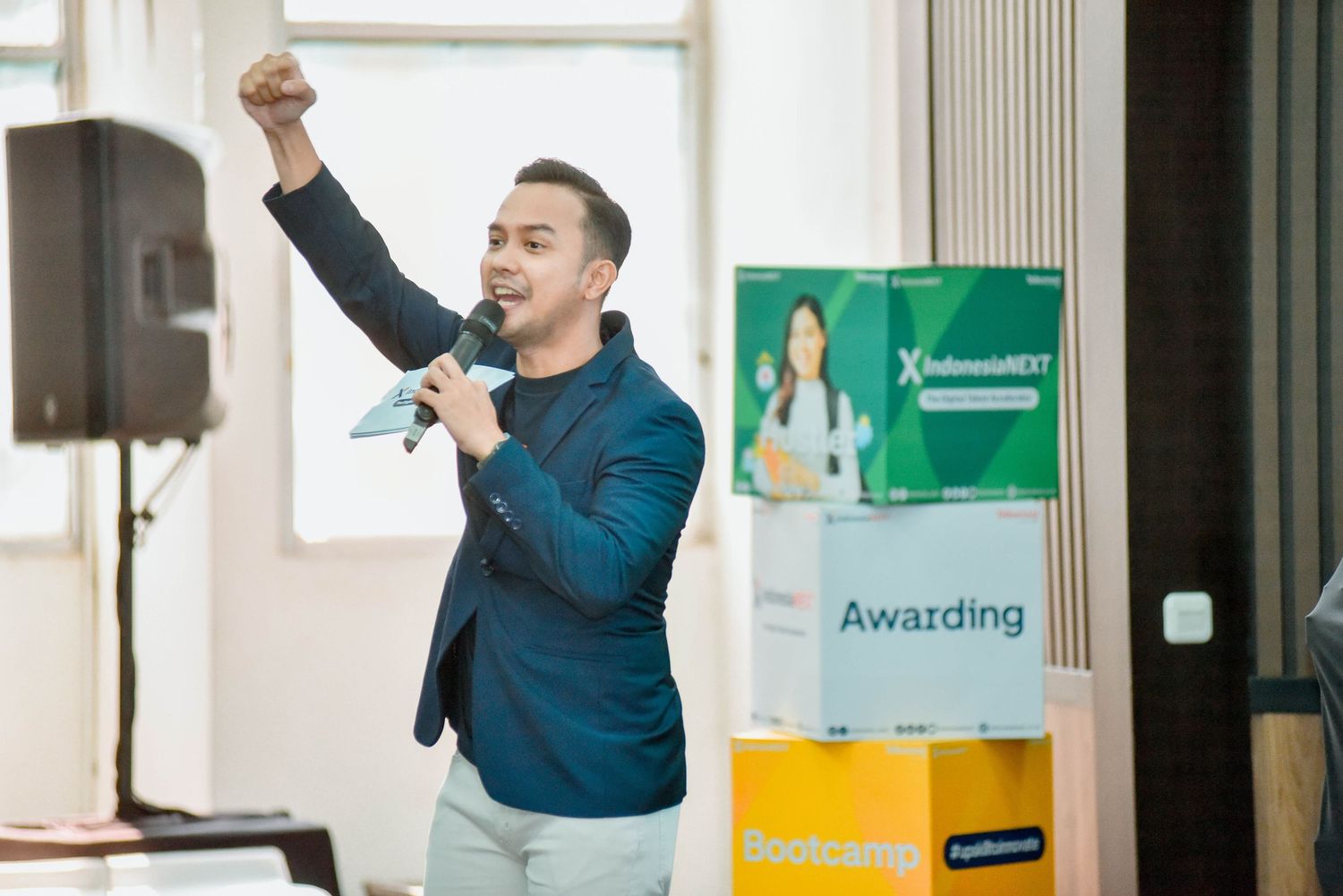 A speaker gestures enthusiastically while presenting at a professional event with colorful banners in the background.
