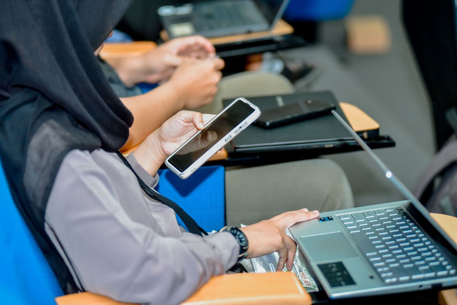 Students work on mobile devices and laptops during a technology class session at a desk with blue chairs.