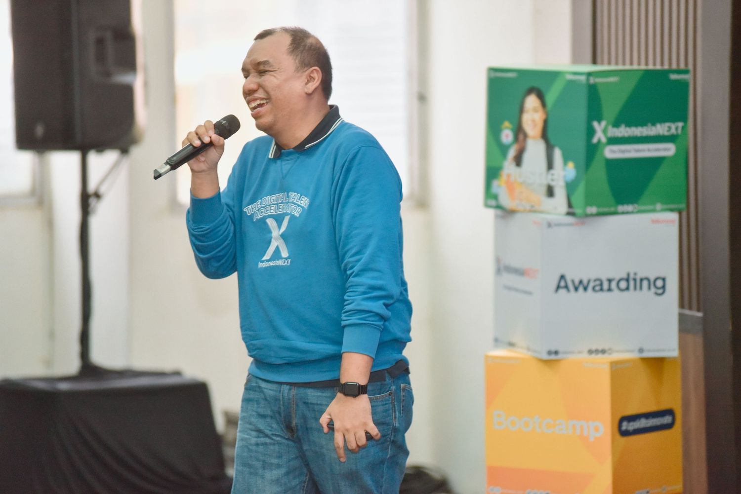 A person in a bright blue sweater speaks into a microphone while gesturing during a presentation.