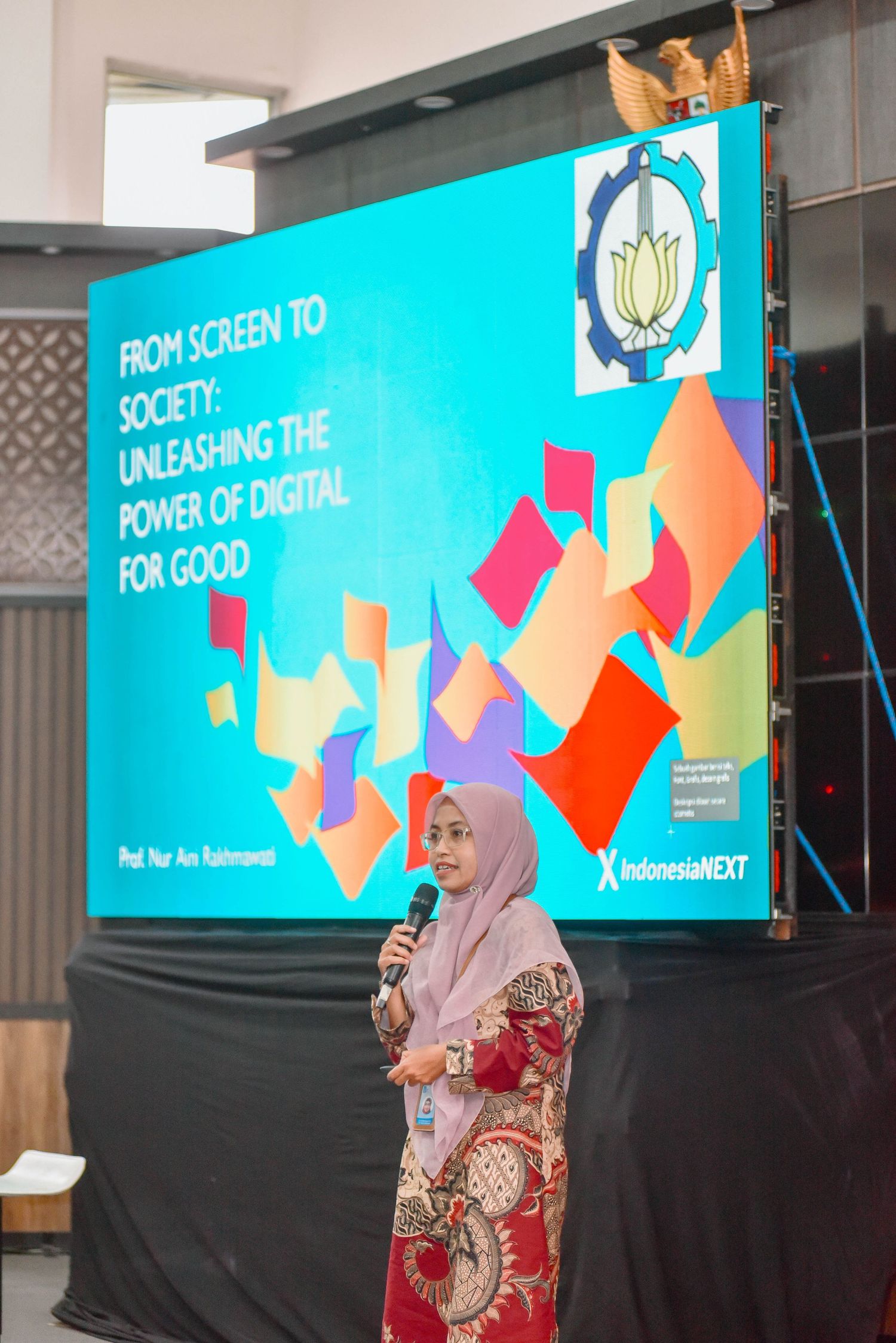 A person in a hijab presents on stage with colorful digital displays showing text and geometric shapes in the background.