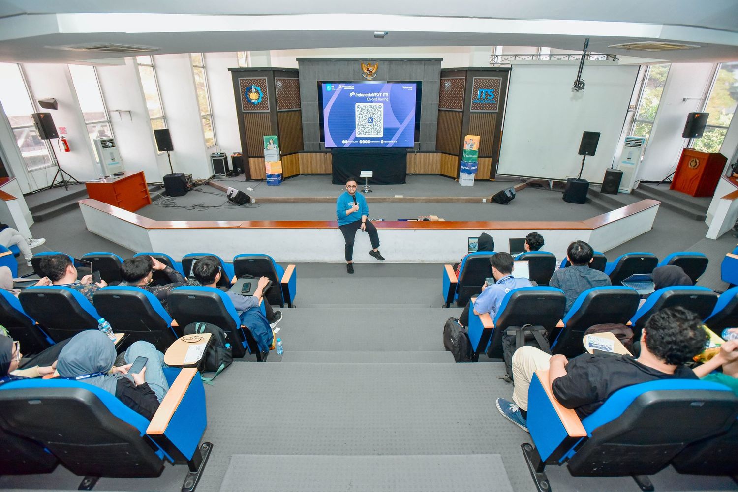 Students in blue uniforms practice martial arts techniques on training mats in a bright indoor facility.