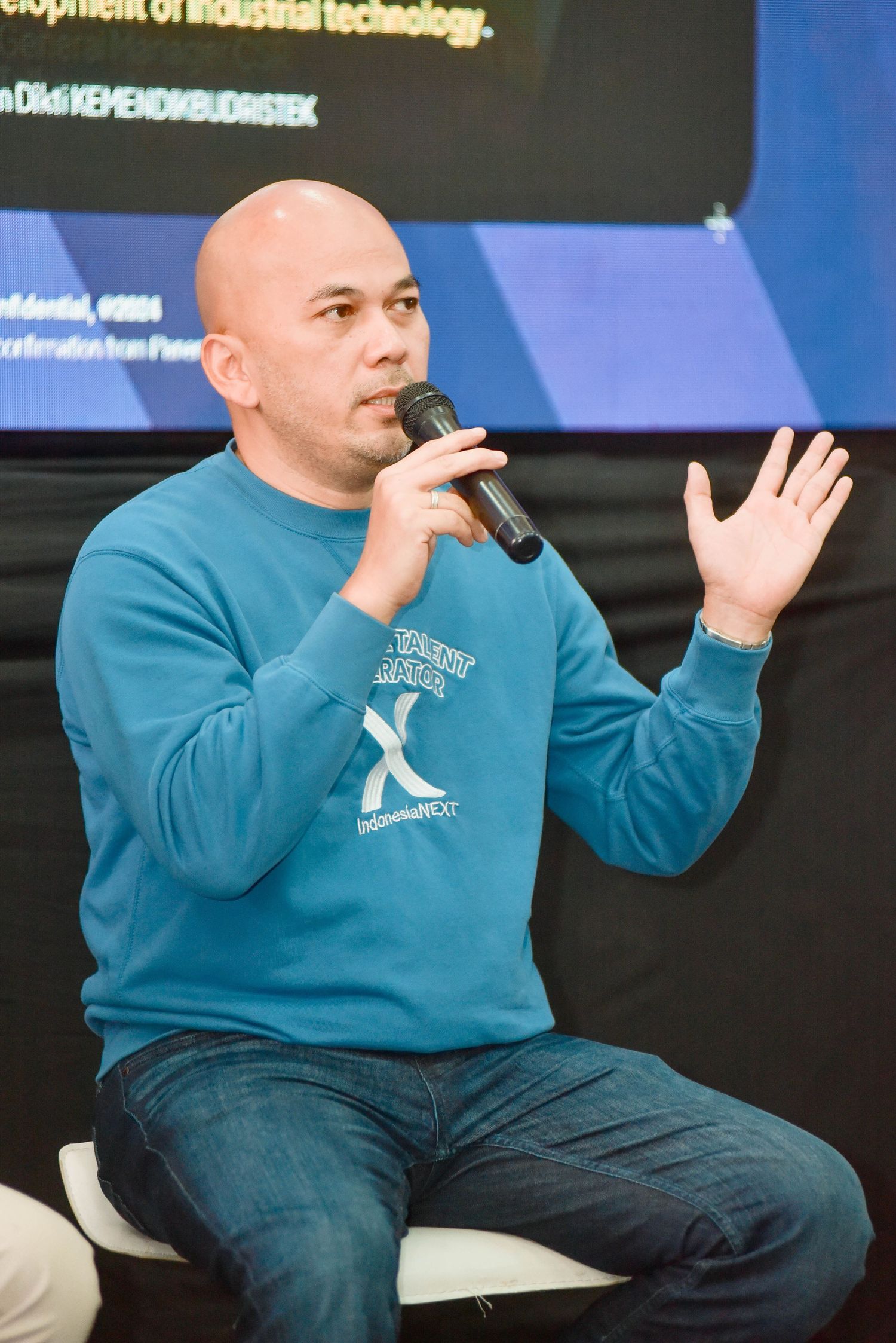 A speaker wearing a teal sweatshirt gestures while holding a microphone during a presentation.