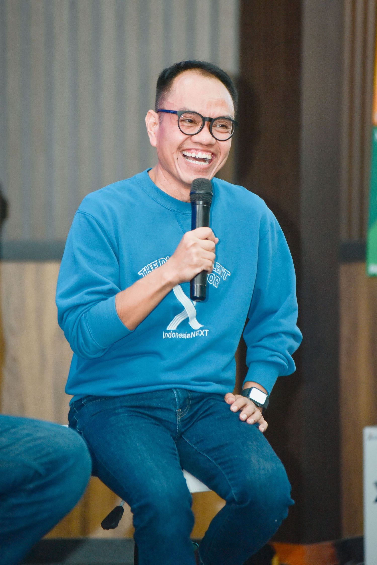 A speaker wearing glasses and a blue sweatshirt sits casually while holding a microphone during a presentation.