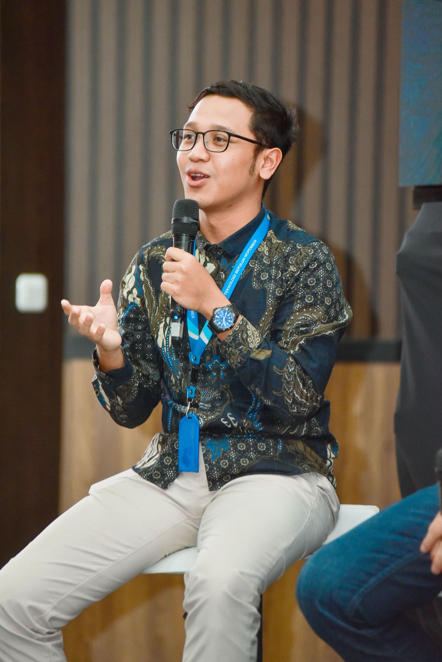 Speaker in a patterned jacket and glasses gestures animatedly during a panel discussion on stage.