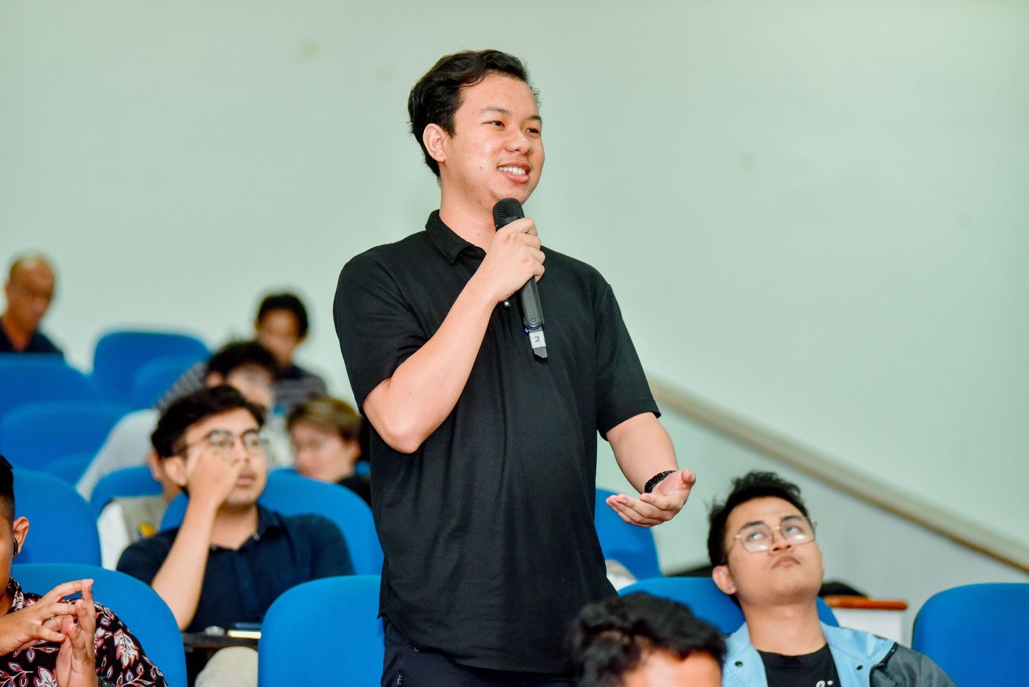 A speaker in a black shirt addresses an audience in blue stadium seating during a presentation.