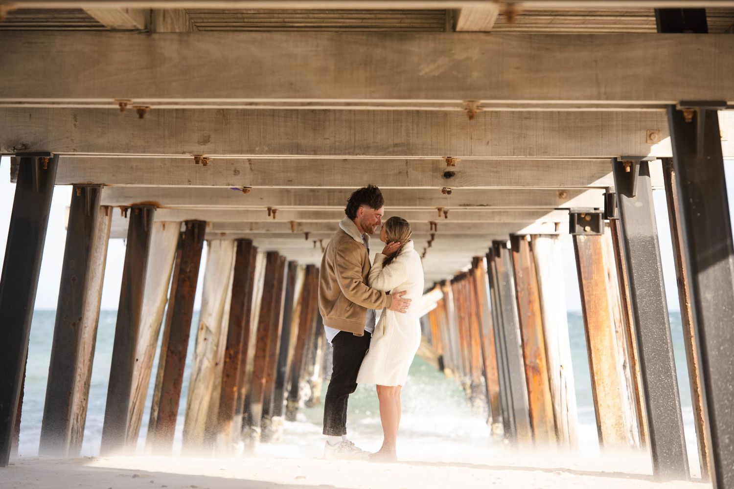 Couple sharing a romantic moment under a pier, surrounded by wooden columns with ocean light in the background.